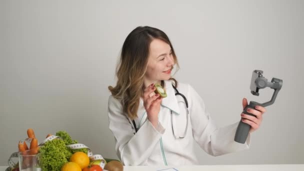 Um médico-nutricionista em sua mesa realiza treinamento on-line para seus clientes por videochamada pelo telefone segurando uma laranja. Alimentação saudável . — Vídeo de Stock