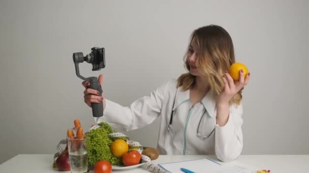 Um médico-nutricionista em sua mesa realiza treinamento on-line para seus clientes por videochamada pelo telefone segurando uma laranja. Alimentação saudável — Vídeo de Stock