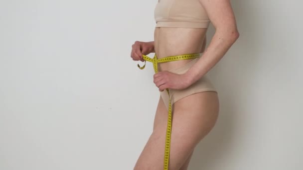 Porträtt av en smal flicka i bruna underkläder som mäter hennes midja med ett måttband efter en diet eller träning och är nöjd med resultatet — Stockvideo