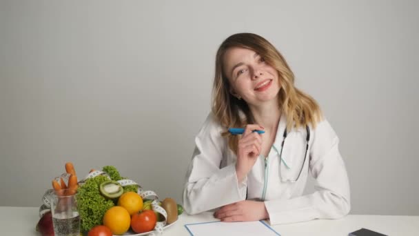 Nutritionist vid sitt skrivbord skriver ett recept för patienten hälsopiller. Grönsaker och frukt på bordet. Lycklig läkare — Stockvideo