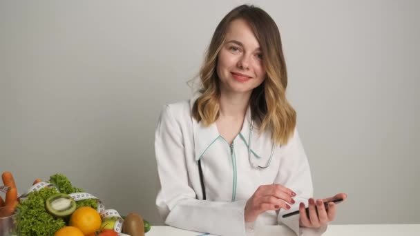 En su escritorio, el nutricionista escribe una receta en el teléfono de los pacientes. Hay píldoras, verduras y frutas en la mesa. Feliz doctor. — Vídeo de stock