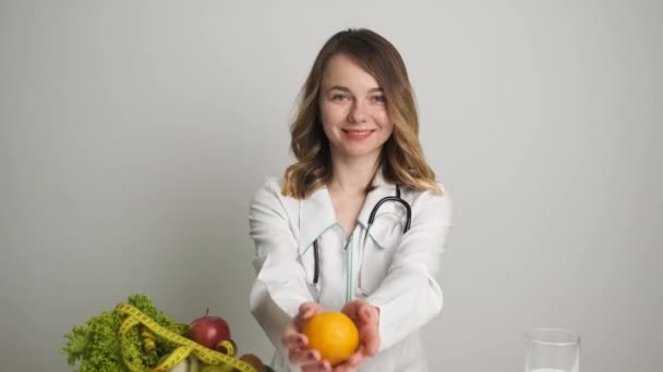 Médica nutricionista com um vestido médico sentado a uma mesa e segurando uma laranja — Vídeo de Stock