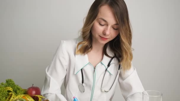 Vrouwelijke arts voedingsdeskundige in een medische jurk zit op het werk en schrijft een gezond dieet — Stockvideo