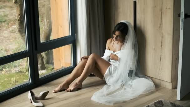 Een mooie bruid in een trouwsluier en badjas zit bij het panoramische raam en groet haar prins. Prachtige bruiloft — Stockvideo