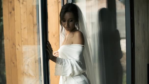 Una bella sposa in un velo nuziale e accappatoio siede vicino alla finestra panoramica e aspetta il suo principe. Bruna incantata con un aspetto caucasico. Bellissimo matrimonio — Video Stock