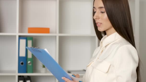 Portrait of a beautiful 25 year old Caucasian looking girl with a sincere smile running standing at her workplace holding a clipboard and checking the contract. Company director — Stock Video