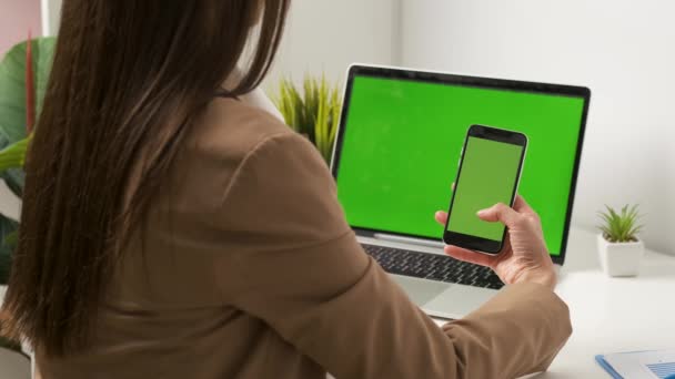Mooie brunette Kaukasische uiterlijk werken in het kantoor op een laptop en kijken naar de telefoon controleert het contract op een groene achtergrond telefoon en laptop. Achterste schot — Stockvideo