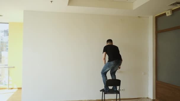 Joven maestro morena hace reparaciones en una gran oficina espaciosa con ventanas panorámicas. Pintar las paredes con un pincel en blanco — Vídeos de Stock