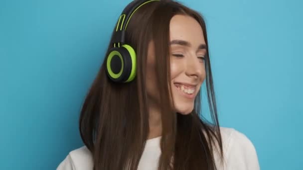 Привлекательная женщина на прочном фоне в стильных зеленых наушниках слушает музыку и улыбается в камеру. Активный образ жизни — стоковое видео