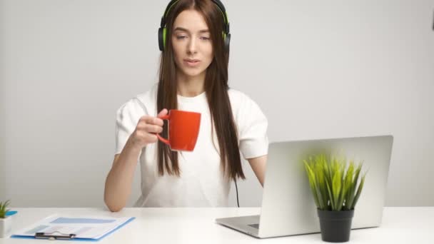 Portré egy vidám könyvelőlányról, aki az irodában ül egy laptopon, zenét hallgat fejhallgatón és kávét iszik. — Stock videók