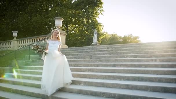 La novia feliz camina en un parque de París. Hermosa rubia baja las escaleras al atardecer en un vestido blanco y con un ramo de flores — Vídeos de Stock