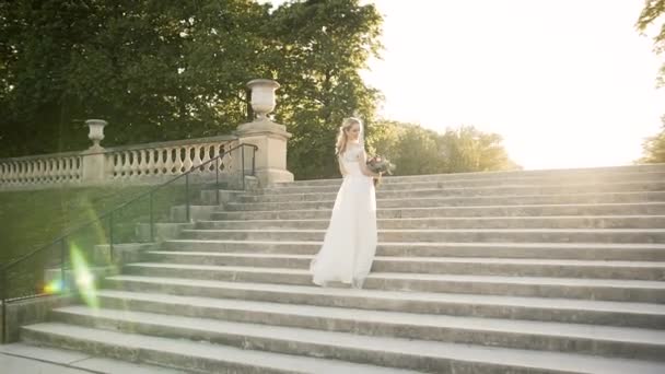 Boldog menyasszony sétál egy parkban Párizsban. Gyönyörű szőke leszáll a lépcsőn naplementekor egy fehér ruhában és egy csokor virággal. — Stock videók