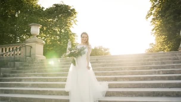 Ευτυχισμένη νύφη περπατάει σε ένα πάρκο στο Παρίσι. Όμορφη ξανθιά κατεβαίνει τις σκάλες στο ηλιοβασίλεμα σε ένα λευκό φόρεμα και με ένα μπουκέτο λουλούδια — Αρχείο Βίντεο