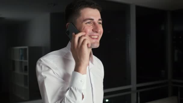 30-jarige zakenman met zwart haar in een stijlvol pak loopt rond het kantoor in de buurt van een panoramisch raam met uitzicht op de nacht stad en praat aan de telefoon bellen met zijn vrouw — Stockvideo