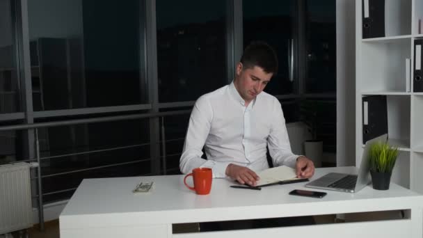 Trzydziestoletni dyrektor firmy siedzi w wieczornym biurze do późna pijąc kawę lub herbatę i czytając książkę. Panorama nocnego miasta z biura — Wideo stockowe