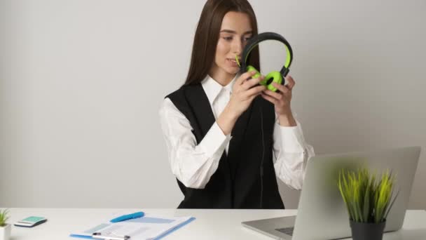Szczęśliwa kobieta w biurze siedzi przy biurku, pracuje na laptopie i słucha muzyki na słuchawkach. — Wideo stockowe