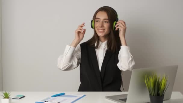 Vidám könyvelő az irodában ül egy asztalnál dolgozik egy laptop és zenét hallgat a fejhallgató — Stock videók