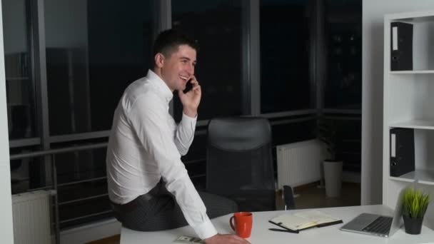 Przystojny mężczyzna zostaje w pracy do późnej nocy, siedzi za biurkiem w biurze z panoramicznymi oknami i telefonem. — Wideo stockowe