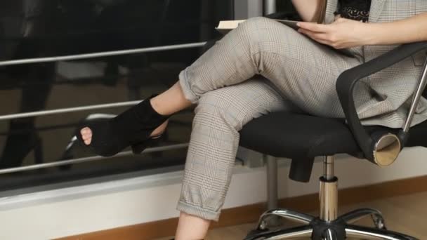 Hermosa chica con el pelo largo sentado junto a la ventana panorámica lee un libro sosteniendo una copa de vino tinto y disfruta de la ciudad nocturna lanzando su pie sobre su pie — Vídeos de Stock