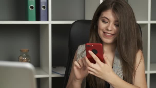 Piękna bizneswoman w garniturze z bujnymi piersiami i szczerym uśmiechem siedzi w biurze w pracy i trzyma telefon w rękach — Wideo stockowe