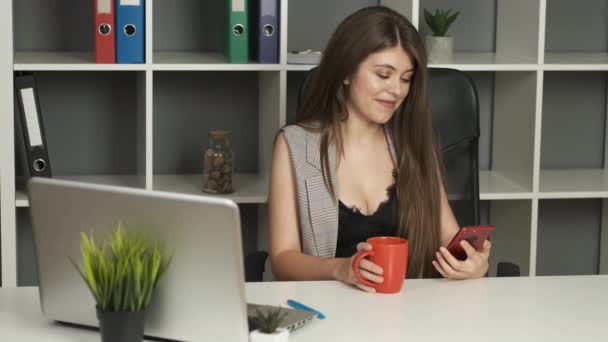 Piękna bizneswoman w garniturze z bujnymi piersiami i szczerym uśmiechem siedzi w biurze w pracy i trzyma telefon w rękach i pije kawę lub herbatę — Wideo stockowe