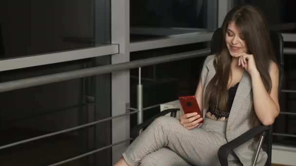 Chica bonita en un traje se sienta en casa en una silla junto a la ventana panorámica y sostiene un teléfono rojo y escribe — Vídeo de stock