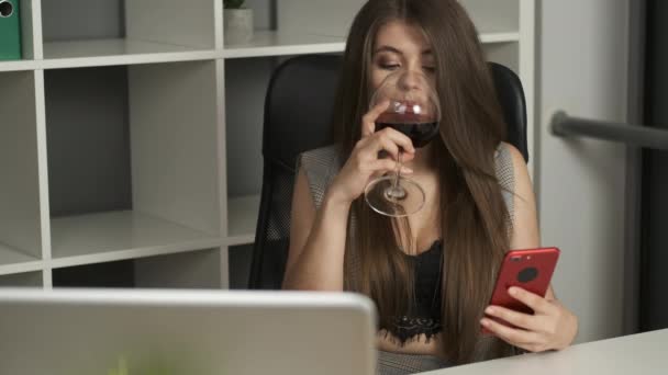 Junge hübsche Firmendirektorin sitzt im Büro, hält ein Telefon in der Hand und trinkt ein Glas Rotwein — Stockvideo
