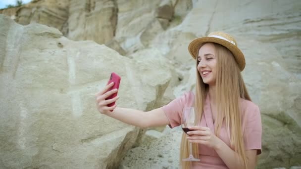 身穿粉色连衣裙、头戴头巾的年轻白种人女游客拿着红色手机，自拍，在峡谷里喝着红酒，欣赏着沙坑的美丽景色 — 图库视频影像