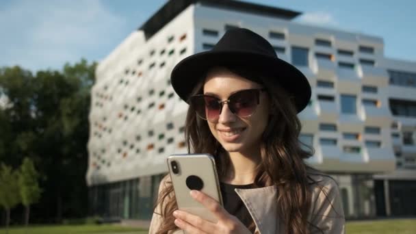 手に電話を持っている近代的な建物の背景には、通りを歩いて誠実な笑顔で眼鏡、レインコート、帽子の若いビジネス女性の肖像画 — ストック動画
