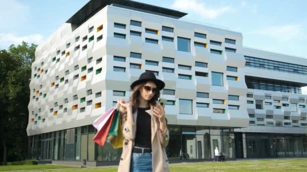 Portret van een jonge zakenvrouw in bril, regenjas en hoed met een oprechte glimlach in het park op de achtergrond van een modern gebouw met een telefoon en cadeauzakjes in haar handen — Stockvideo