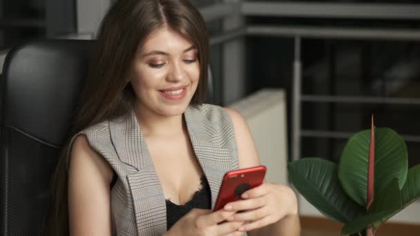 Ładna dziewczyna w garniturze siedzi w domu na krześle przy panoramicznym oknie i trzyma czerwony telefon i pisze — Wideo stockowe