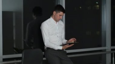 Beyaz gömlekli beyaz tenli esmer, panoramik pencerenin yanında duruyor. Gece şehrine bakıyor ve bir kitap okuyor.