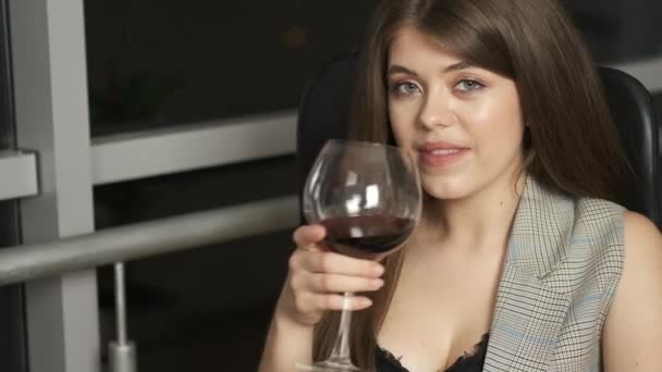 Gadis cantik dari penampilan Kaukasia dengan rambut panjang duduk di jendela panorama membaca sebuah buku memegang segelas anggur merah dan menikmati kota malam — Stok Video
