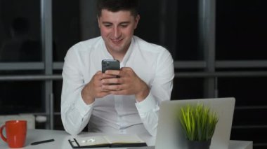 Yakışıklı esmer, geceleri ofiste takılıyor, elinde telefon ve kulaklıkla dizüstü bilgisayarla müzik dinliyor.