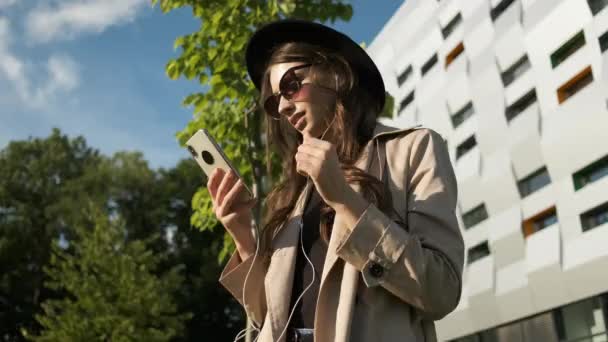 Portret van een jonge zakenvrouw in bril, regenjas en hoed met een oprechte glimlach loopt door de straat op de achtergrond van een modern gebouw met een telefoon in zijn handen — Stockvideo