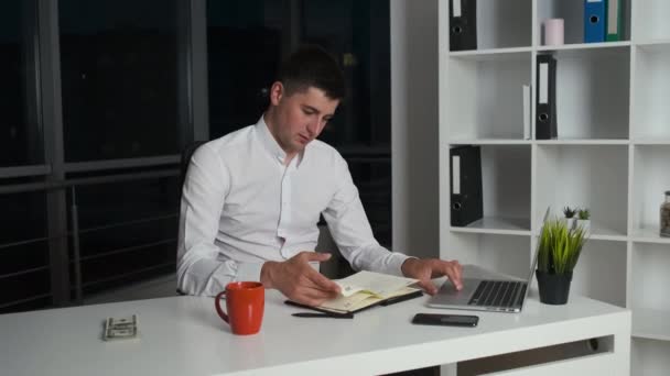 Le directeur de l'entreprise, âgé de 30 ans, est assis dans le bureau du soir jusqu'à ce qu'il boive tardivement du café ou du thé et lise un livre. Panorama de la ville nocturne depuis le bureau — Video