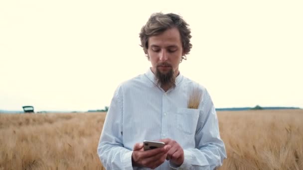 Een jonge man met een baard loopt op een tarweveld en neemt gegevens op over de telefoon. — Stockvideo