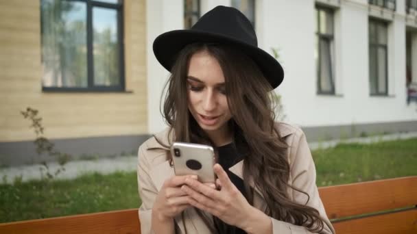 Portrait d'une jeune femme d'affaires en lunettes, imperméable et chapeau avec un sourire sincère assise sur un banc sur le fond d'un bâtiment moderne tenant un téléphone — Video