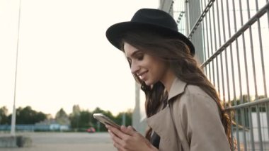 Sokakta elinde telefonla bekleyen güzel bir kız. Esmer, uzun saçlı, siyah şapkalı ve kahverengi pelerinli.