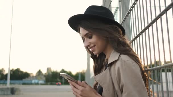 Красивая девушка, стоящая на улице с телефоном. Брюнетка с длинными волосами в черной шляпе и коричневом плаще — стоковое видео