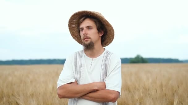 Een brutale man met een baard en een strohoed staat in een tarweveld met gevouwen handen. Zomer zonnige avond — Stockvideo