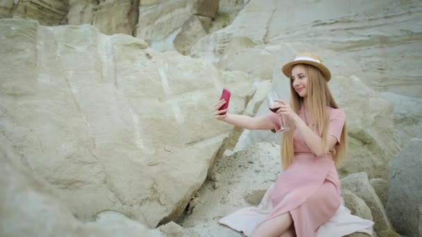 Туристка молодая кавказская блондинка в розовом платье с капюшоном держит красный телефон и делает селфи и выпивает бокал красного вина в каньоне с прекрасным видом на пещеру — стоковое видео