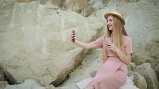 Туристка молодая кавказская блондинка в розовом платье с капюшоном держит красный телефон и делает селфи и выпивает бокал красного вина в каньоне с прекрасным видом на пещеру — стоковое видео