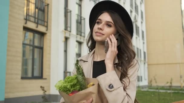 Gyönyörű, elegáns nő barna köpenyben és fekete kalapban sétál az utcán a modern épületek közelében, kezében egy csomag zöldséggel és gyümölccsel, és telefonon beszél. — Stock videók