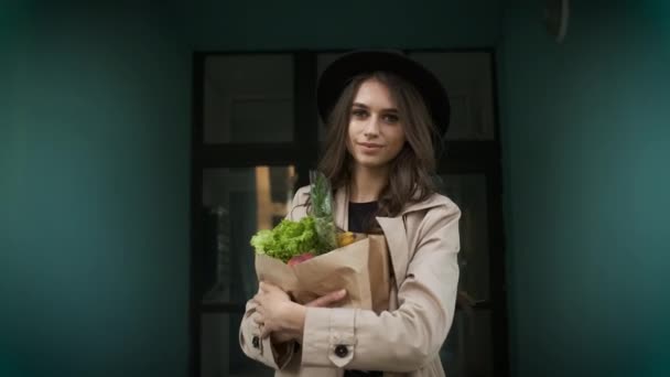 Hermosa mujer con estilo en una capa marrón y sombrero negro camina por la calle cerca de edificios modernos, sosteniendo un paquete de verduras y frutas y un teléfono en sus manos. Entrega de comida a casa — Vídeo de stock