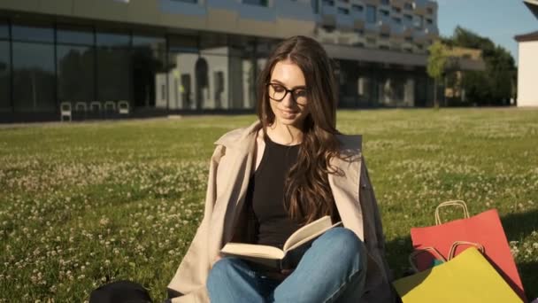 Gözlüklü, kahverengi pelerinli şık bir kız parkta çimlerin üzerinde oturur ve kitap okur. Hediye paketleri. Yaz güneşli bir akşam — Stok video