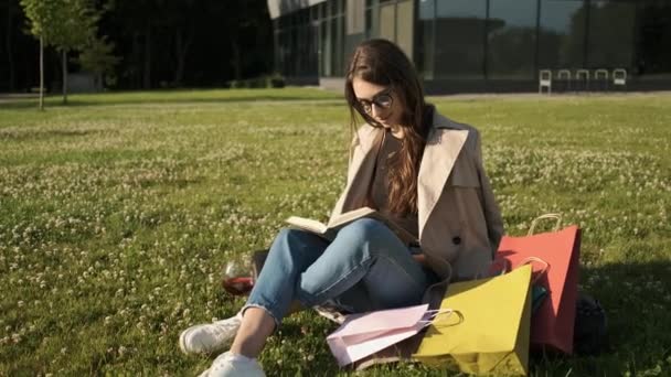 Ragazza elegante con bicchieri e un mantello marrone si siede nel parco sull'erba e legge un libro e beve un bicchiere di vino rosso. Pacchetti regalo. Serata estiva soleggiata — Video Stock