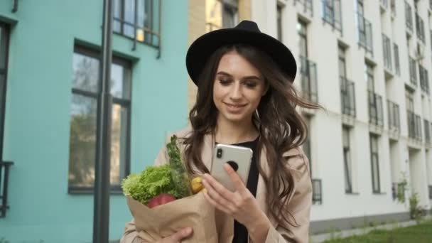 Belle femme élégante dans un manteau brun et chapeau noir marche dans la rue près des bâtiments modernes, tenant un paquet de légumes et de fruits et un téléphone dans ses mains. Livraison de nourriture — Video