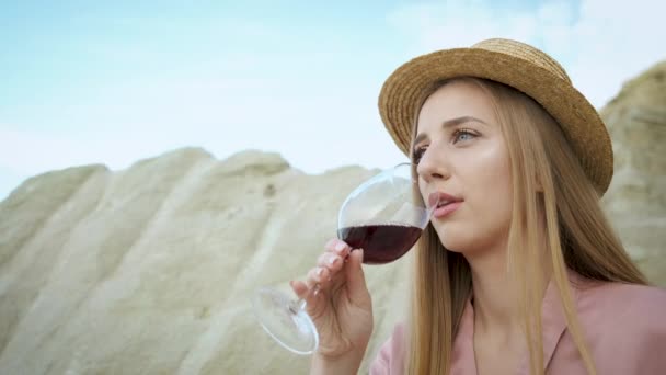 Ragazza di 25 anni turista in un cappello biondo aspetto caucasico siede su una pietra in una cava di sabbia e beve un bicchiere di vino rosso e si rilassa. Godersi il canyon. — Video Stock