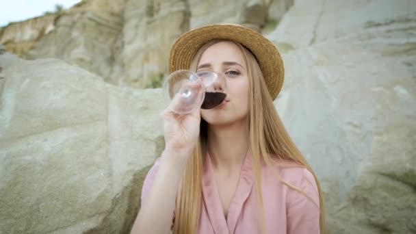 25 jaar oud meisje toerist met een blonde hoed Kaukasische verschijning zit op een steen in een zandgroeve en drinkt een glas rode wijn en ontspant. Genieten van de canyon — Stockvideo